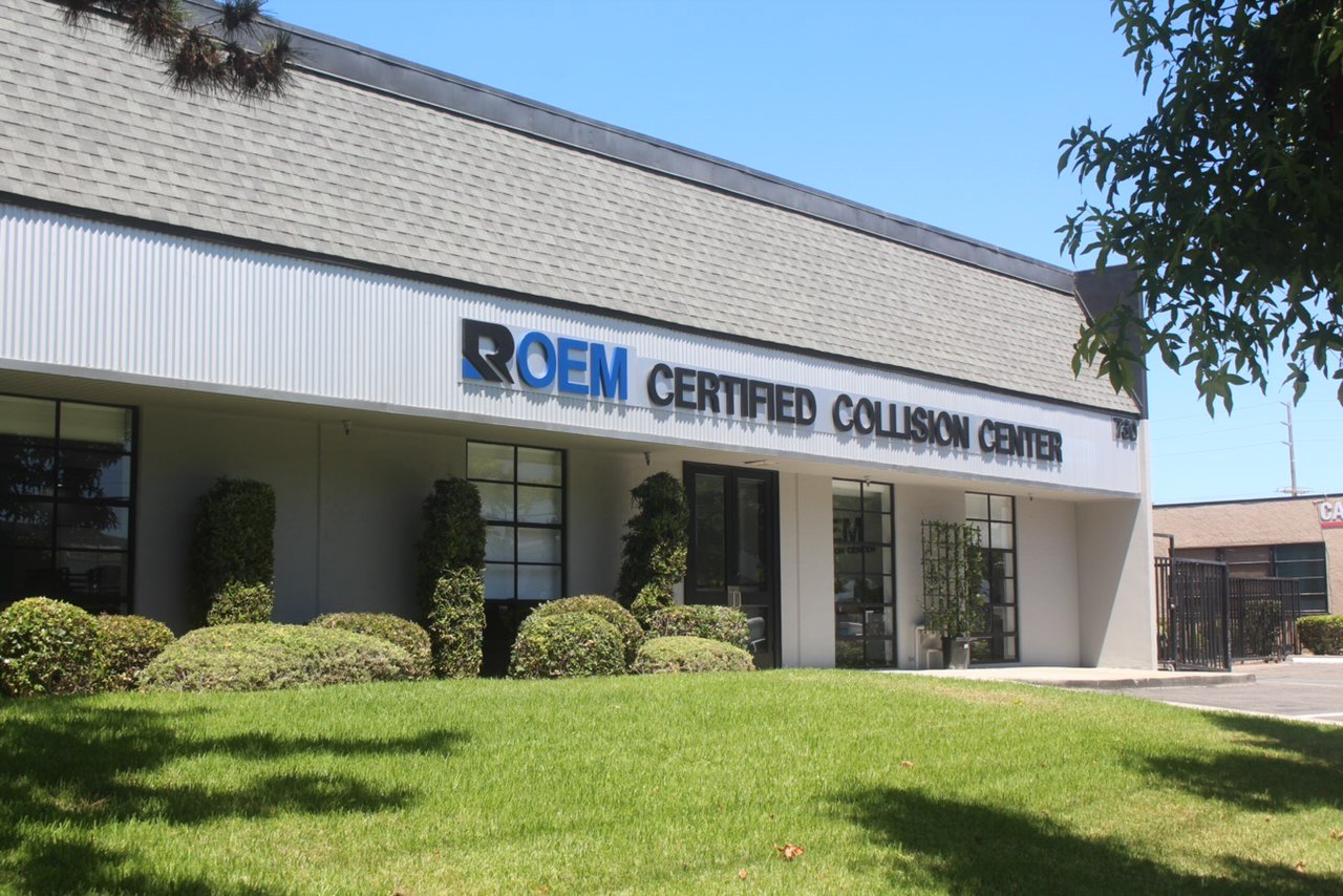 Commercial building with a “ROEM CERTIFIED COLLISION CENTER” sign, landscaped shrubs, and a sunny lawn.